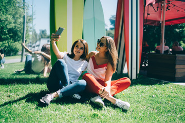 Carefree trendy millennial women taking selfie with smartphone while chilling together in park