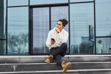 Handsome young man with curly black hair and with smartphone in hands is on the street sitting on the stairs against building