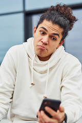 Handsome young man with curly black hair sitting on the street with smartphone against building