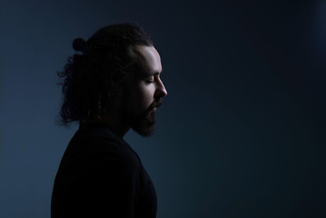 studio dramatic profile portrait of a young bearded guy of thirty years old, with long hair gathered, looks down. On a dark background. A ray of light on the face. Dramatic light
