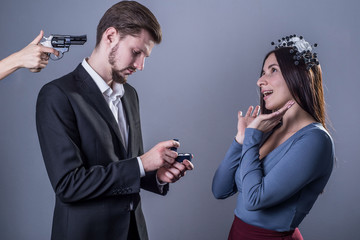 Studio portrait of a sad guy on his knees proposing marriage to a happy girl standing next to him, he faces a pistol from behind. On a gray background. wedding calculation concept.