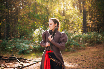 A girl stands in a dark forest, in a brown coat with a red belt and boots. Slovenian girl.