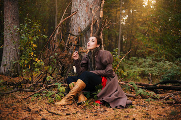 Naklejka na ściany i meble A girl sits under a tree in a dark forest, in a brown coat and boots. Slovenian girl chewing a blade of grass.