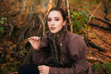 A girl sits under a tree in a dark forest, in a brown coat and boots. Slovenian girl chewing a...