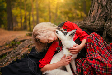 A girl in a plaid skirt, a red sweater and a raincoat sits by a tree and hugs a white fox. The blonde in the forest in the image of a peasant woman