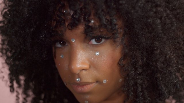 Mixed race black woman with curly hair covered by crystal makeup on pink background in studio Close up show ing makeup