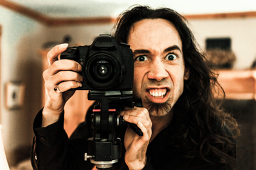 A close up front portrait of a caucasian photographer with a funny creepy face expression, standing behind a professional DSLR camera and tripod.