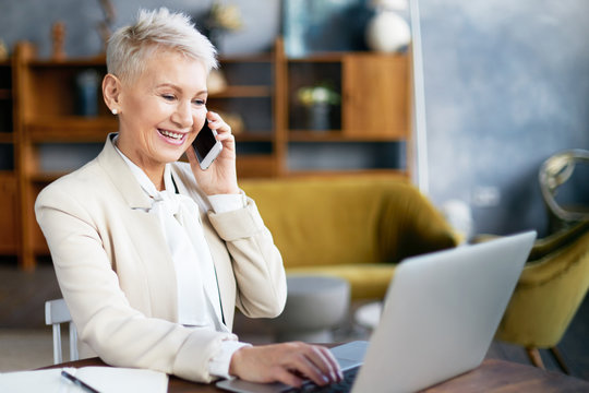 Indoor Shot Of Attractive Mature Female Sales Manager In Stylish Suit Having Phone Conversation Using Mobile, Selling Goods Or Services Online Via Internet, Sitting At Desk In Front Of Open Laptop