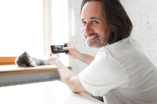An adult man takes pictures on the phone of his cat.
