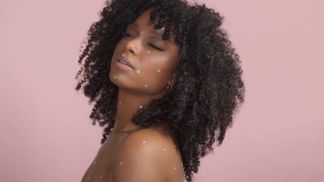 Mixed race black woman with curly hair covered by crystal makeup on pink background in studio Model watching back and dancing