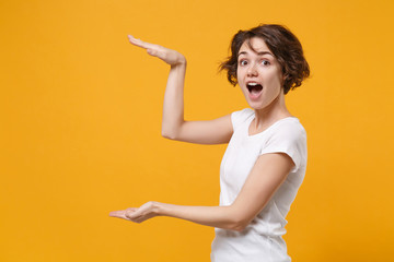 Shocked young brunette woman in white t-shirt posing isolated on yellow orange background in studio. People lifestyle concept. Mock up copy space. Gesturing demonstrating size with vertical workspace.