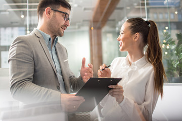 Businessman and businesswoman in formalwear discussing something while holding clipboard with...