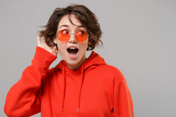 Shocked young brunette woman girl in casual red hoodie eyeglasses posing isolated on grey...