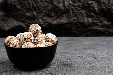 Homemade energy balls of dates, peanuts, oats, sprinkled with coconut in a black bowl on a dark background with copy space with place for text.