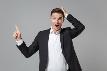 Excited young business man in classic black suit shirt isolated on grey wall background. Achievement career wealth business concept. Mock up copy space. Pointing index finger up, put hand on head.