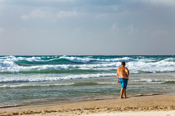 Fat man with a large stomach walking on the beach and looking to stormy sea. Health crisis concept.