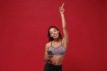 Fototapeta na wymiar Young african american sports fitness woman in sportswear posing working out isolated on red background. Sport exercises healthy lifestyle concept. Hold mobile phone listen music with earpods dancing.
