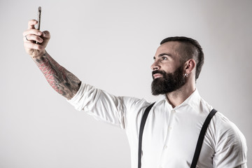 Uomo con barba camicia bianca e bretelle nere,  utilizza il suo smartphone per farsi un selfie,...