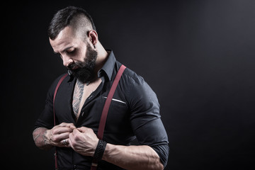Ragazzo barbuto con Tatuaggi in tutto il corpo, vestito con camicia nera e bretelle rosse, si aggancia un bottone, isolato su sfondo nero