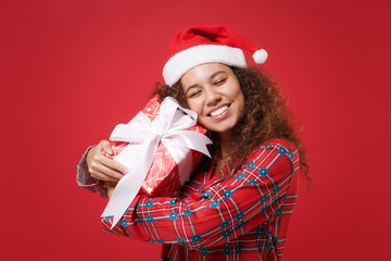 Pretty african american girl in pajamas homewear Christmas hat rest at home isolated on red background. Relax good mood lifestyle concept. Hold present box with gift ribbon bow keeping eyes closed.