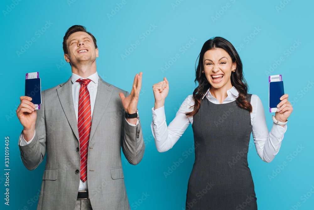 Wall mural two excited businesspeople showing winner gestures while holding passports and air tickets on blue background