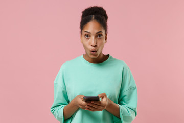 Shocked young african american woman girl in green sweatshirt posing isolated on pastel pink background in studio. People lifestyle concept. Mock up copy space. Using mobile phone, typing sms message.