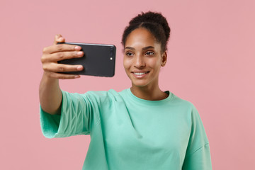 Smiling young african american woman girl in green sweatshirt posing isolated on pastel pink wall background in studio. People lifestyle concept. Mock up copy space. Doing selfie shot on mobile phone.