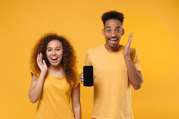 Excited friends couple african american guy girl in casual clothes isolated on yellow background. People lifestyle concept. Mock up copy space. Spreading hands, hold mobile phone with empty screen.