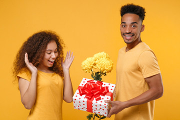 Side view of excited friends couple african american guy girl in casual clothes isolated on yellow background. People lifestyle concept. Hold bouquet of flowers, red present box with gift ribbon bow.