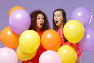 Fototapeta na wymiar Shocked two european african american women friends in knitted sweaters isolated on violet purple background. Birthday holiday party, people emotions concept. Celebrating hold colorful air balloons.