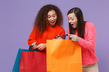 Shocked two young european african american women friends in knitted sweaters isolated on violet purple background in studio. People lifestyle concept. Hold package bag with purchases after shopping.