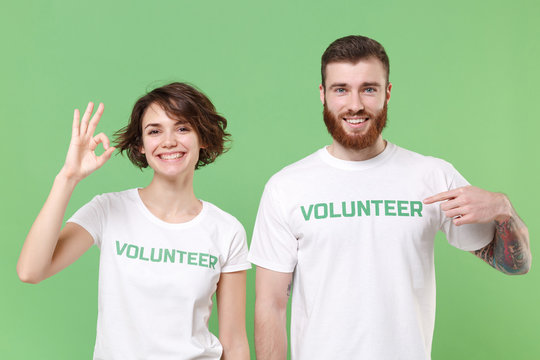 Funny two young friends couple in white volunteer t-shirt isolated on pastel green background studio portrait. Voluntary free work assistance help charity grace teamwork concept. Showing OK gesture.