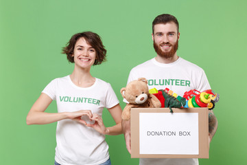 Smiling friends couple in volunteer t-shirt isolated on green background. Voluntary free work assistance help charity grace teamwork. Hold donation box with kids toys, showing shape heart with hands.