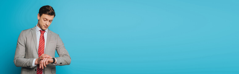 handsome, serious businessman checking time on wristwatch on blue background
