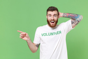 Surprised young bearded man in white volunteer t-shirt isolated on green background. Voluntary free...