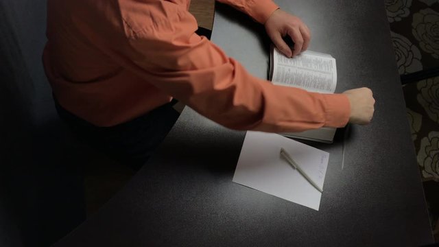 A man reads the Bible while sitting at the table. Writes important thoughts on a piece of paper. Corrects a bookmark and closes the book.