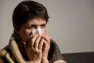  A sick older woman sneezes into a napkin, and covered in a blanket at home. An ill woman sitting on the sofa alone.  Coronavirus. COVID-19. A quarantine at home. 