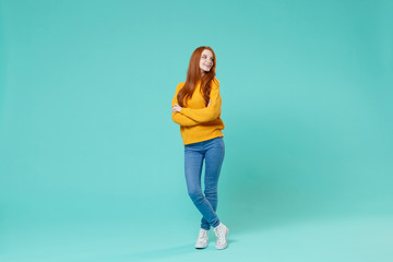Pretty young redhead woman girl in yellow sweater posing isolated on blue turquoise wall background studio portrait. People lifestyle concept. Mock up copy space. Holding hands crossed, looking aside.