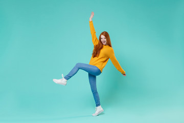 Fototapeta na wymiar Cheerful young redhead woman girl in yellow sweater posing isolated on blue turquoise background studio portrait. People lifestyle concept. Mock up copy space. Rising hand and leg having fun, dancing.