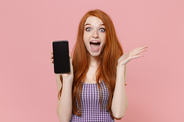 Surprised young redhead woman girl in plaid dress posing isolated on pastel pink background. People lifestyle concept. Mock up copy space. Hold mobile phone with blank empty screen spreading hands.