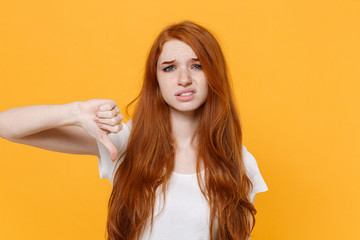 Displeased young redhead woman girl in white blank empty t-shirt posing isolated on yellow wall background studio portrait. People emotions lifestyle concept. Mock up copy space. Showing thumb down.