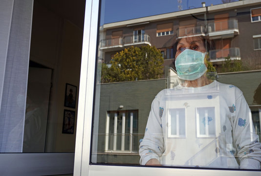 Europe, Italy, Milan - Pandemic Emergency N-cov19 Coronavirus - Domestic Life In Quarantine Of 70 Year Old Lady Who Carries Out Various Activities At Home With The Mask Looking Outside From The Window