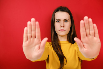 Beautiful serious young brunette woman pulls hands towards camera in stop gesture, asks to calm down and stop worrying, shows limit, wears yellow sweater, isolated over on red background.