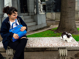 Young woman sitting on a bench looking at a cat that lives in a cemetery. La Recoleta Cemetery is a cemetery located in the Recoleta neighbourhood of Buenos Aires.