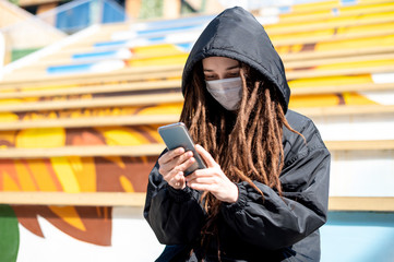 girl with dreadlocks in a medical mask looks at a smartphone