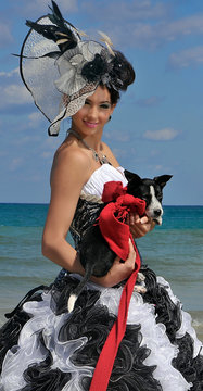 Quinceañera posando elegantemente en la playa con perro en las manos y sombrero y vestido blanco y negro con fondo azul de plata y arena en Cuba, La Habana