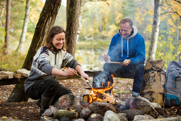 Man and woman making pancakes on campfire in forest on shore of lake, making a fire, grilling. Happy couple exploring Finland. Scandinavian landscape. 