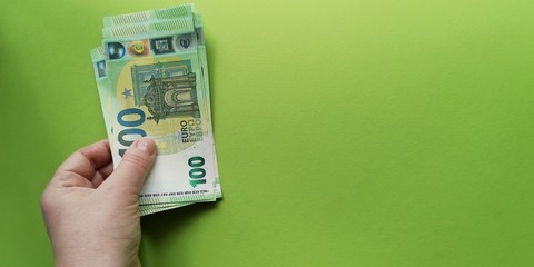 Left hand holding stack of Euro banknotes issued in 2019 with face value of one hundred on green backdrop.Concept of limiting cash payments to prevent the spread of coronavirus.Soft focus, copy space.