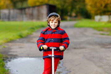 cute little school kid boy riding on push scooter on the way to or from school. Schoolboy of 7 years driving through rain puddle. funny happy child in colorful fashion clothes and with helmet.