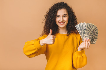 Portrait of a cheerful young curly woman holding money banknotes and celebrating isolated over beige background. Thumbs up.